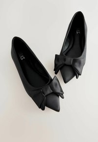 Ballerines en satin noir avec des pointes aigües, présentant un grand nœud à l'avant. Texture lisse et design minimaliste. Légères et élégantes.