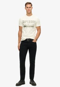 Hvid bomulds t-shirt med sort "SUPERDRY SURPLUS NEW YORK" print, parret med sorte slim-fit jeans og sorte snørebåndssko.