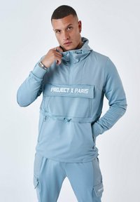 Ljusblå huvtröja med hög krage, dragsko-detalj och framficka. Har vit text "PROJECT X PARIS".