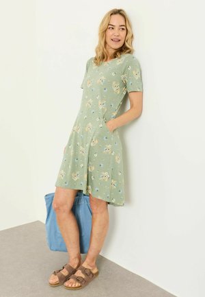 Femme en robe verte à fleurs à manches courtes, les mains dans les poches, sandales marron et un grand sac bleu posé à côté d'elle sur le sol.