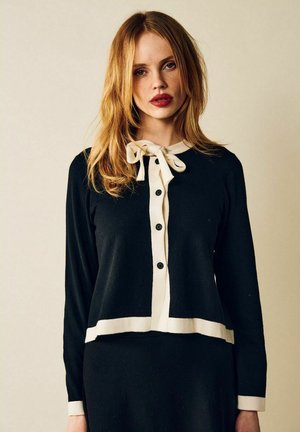Musta neule Cardigan, jossa on kontrastinen valkoinen kaulus ja nappilista. Pitkät hihat ja lyhennetty malli. Rusettiakseli lisää yksityiskohtia.
