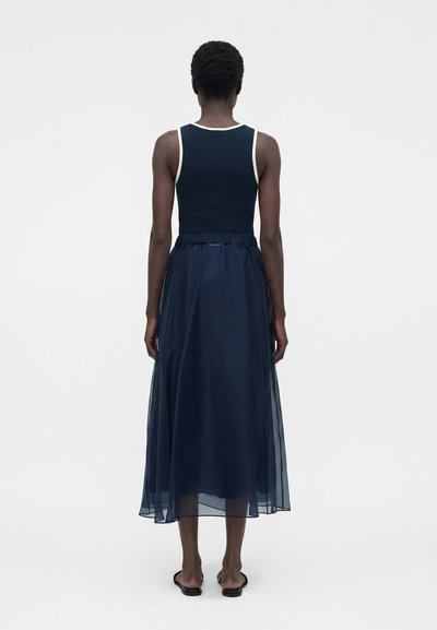 Άτομο με κοντά σγουρά μαλλιά που φοράει ένα μαύρο τοπ χωρίς μανίκια και μια διαφανή μπλε-σκούρα μίντι φούστα, στέκεται σε ένα απλό λευκό φόντο.