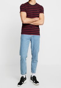 Camiseta de manga corta a rayas negras y rojas, corte casual, combinada con jeans de mezclilla azul claro y zapatillas negras con detalles en blanco.