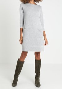 Donna che indossa un vestito grigio chiaro fino al ginocchio con maniche a tre quarti e grandi tasche frontali, abbinato a stivali in suede verde scuro al ginocchio.