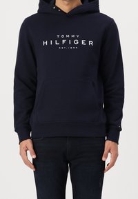 Uomo che indossa un hoodie navy blue di Tommy Hilfiger con tasca frontale e testo del logo bianco, abbinato a jeans blu scuro.