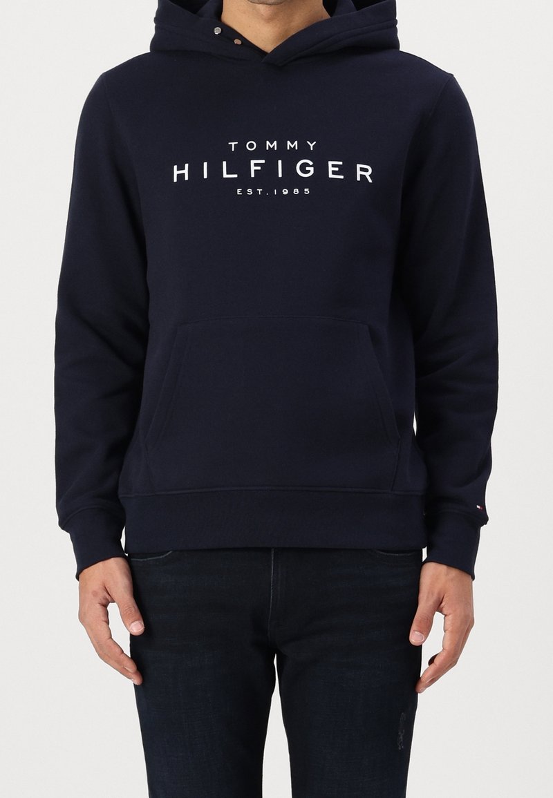 Uomo che indossa un hoodie navy blue di Tommy Hilfiger con tasca frontale e testo del logo bianco, abbinato a jeans blu scuro.