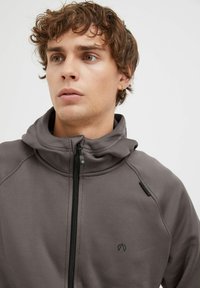 Giacca grigia con zip e colletto alto, caratterizzata da una superficie liscia e dettagli delle cuciture. Un piccolo logo è visibile sul lato sinistro.