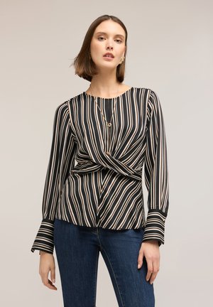 Žena oblečená v černobélo-béžovém pruhovaném blůze s dlouhým rukávem, která je u pasu zauzlená, tmavě modrých džínách a zlatých špercích.