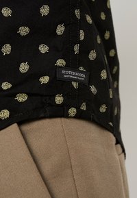 Camicia nera con motivo a foglia d'oro, caratterizzata da un'etichetta che recita "SCOTCH & SODA", abbinata a pantaloni beige con un tessuto testurizzato.