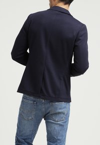 Navy blazer with a smooth texture, tailored design, and single vent at the back, paired with blue denim jeans featuring back pockets.