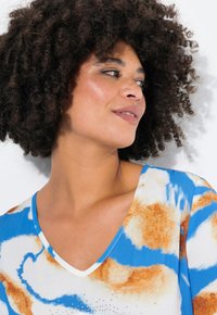 T-shirt a V con motivo tie-dye blu e arancione, realizzato in tessuto morbido e con una leggera lucentezza. Decorato con piccole borchie vicino al collo.