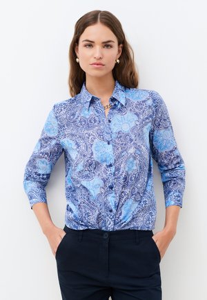 Mujer con cabello largo castaño, que lleva pendientes dorados y una camisa azul con estampado floral paisley abotonada, metida dentro de pantalones oscuros, de pie con las manos en los bolsillos.