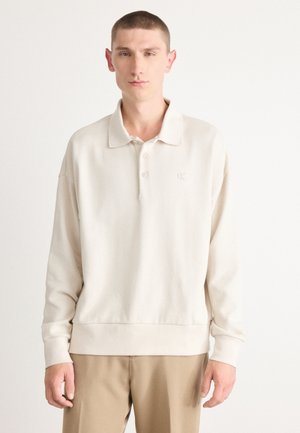 Beige pitkähihaine polo-paita pehmeällä pinnalla, kolmella napilla varustettu liepe, ja huomaamaton 'CK' logo brodeerattuna rintaan. Yksinkertainen muotoilu.