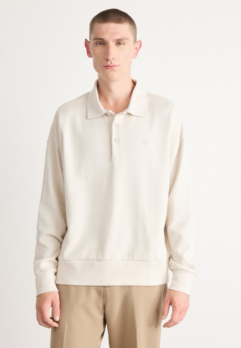 Beige Langarm-Polohemd mit weicher Textur, drei-Knopf-Leiste und dezent eingesticktem 'CK'-Logo auf der Brust. Schlichtes Design.