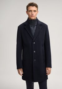 Cappotto di lana blu scuro con un design su misura, dotato di colletto a rever, quattro bottoni e tasche laterali, indossato sopra un gilet imbottito nero.