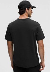 Camiseta negra de manga corta con cuello redondo. Tejido suave, corte relajado, llevada con una gorra negra a juego, vista desde un ángulo trasero.