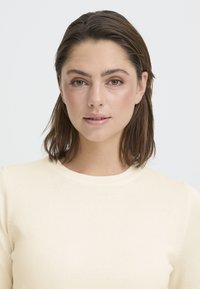 Jeune femme aux cheveux bruns mi-longs, yeux verts et maquillage naturel portant un chemisier côtelé de couleur crème, fond neutre.