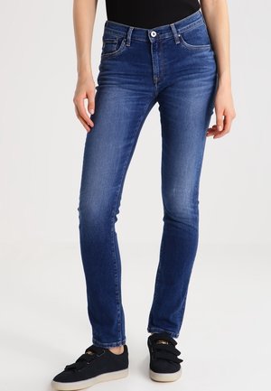 Femme portant un jean slim bleu et des baskets noires, debout devant un fond clair uni.