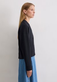 Cardigan en coton noir avec un col rond et une fermeture à boutons, associé à une jupe en denim bleu clair, présenté en vue de profil.