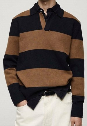 Mand iført en sort og brun bredstribet langærmet polotrøje over en sort skjorte med krave, med hvide bukser og et sort bælte.