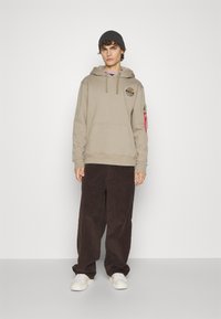 Felpa beige con logo NASA, tasca frontale e stringhe; abbinata a pantaloni in corduroy marroni e sneakers bianche. Modello che indossa un berretto grigio.