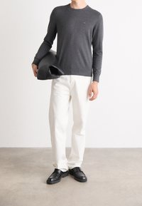 Homme portant un pull à manches longues gris foncé, pantalon blanc, chaussures noires, tenant une veste en cuir noire pliée dans un cadre intérieur minimaliste.