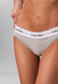 Gros plan sur une personne portant un slip Calvin Klein gris clair avec une ceinture blanche portant le texte noir Calvin Klein.