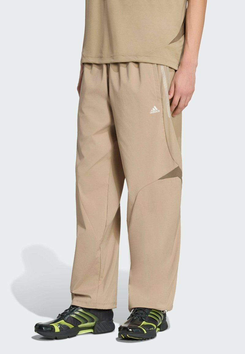 Pantalon de sport beige à coupe ample, avec des accents texturés et un design ventilé. Associé à des chaussures de sport noires et vertes.
