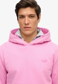 Felpa con cappuccio rosa dal texture morbido. Presenta un logo ricamato sul petto e un colletto a strisce stratificato sotto.