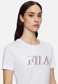 Witte katoenen T-shirt met korte mouwen, voorzien van een groot, gestileerd "FILA" logo in rood en grijs, centraal op de borst geplaatst.
