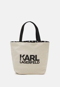 Sac fourre-tout en toile avec poignées noires, extérieur crème et intérieur noir, orné d'une impression de dessin animé. L'avant affiche le logo "KARL LAGERFELD" en gras noir.