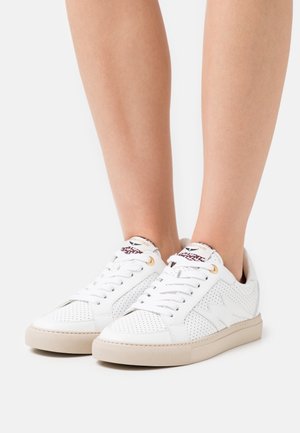Sneakers in pelle bianca traforata con suole beige indossate a piedi nudi, con lacci e un delicato motivo a stella vicino ai lati.