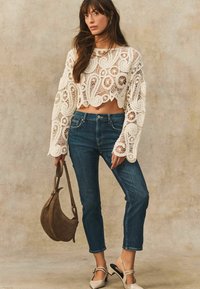 Biały, dziergany sweter w kroju crop z misternymi wzorami, zestawiony z ciemnoniebieskimi dżinsami i beżowymi pantoflami na klamrze. Brązowa skórzana torba na ramię.