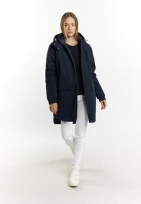 Mørkeblå lang puffjakke med hætte, to forlommer, elastiske manchetter og fuld lynlås, båret over en mørk top og hvide bukser.