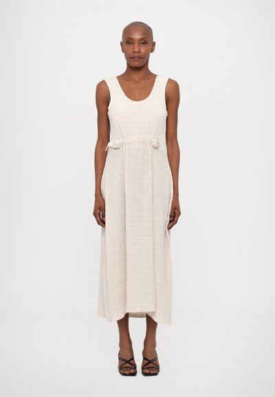 Robe maxi sans manches beige en tissu texturé, cintrée à la taille avec des cordons à nouer et dotée de poches latérales. Sandales noires à lanières.
