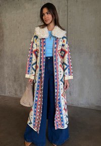 Γυναίκα που φορά μακρύ παλτό με σχέδια και γιακά από shearling, ανοιχτό μπλε τοπ, τζιν με φαρδιά μπατζάκια, καφέ παπούτσια, κρατώντας μια μπεζ τσάντα μπροστά από έναν τοίχο από σκυρόδεμα.