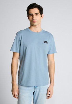 Mężczyzna ubrany w jasnoniebieską koszulkę z krótkim rękawem z małą czarną naszywką z logo na piersi oraz jasne dżinsy, stojący na jednolitym tle.