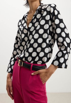 Vrouw draagt een zwart-wit polkadot blouse, gestopt in een hoog uitgesneden roze broek met een zwarte riem en een gouden gesp.