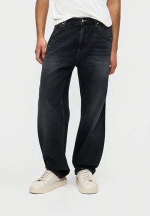 Homme portant un jean en denim noir ample avec des détails délavés, un t-shirt blanc et des baskets blanches, debout devant un fond uni.