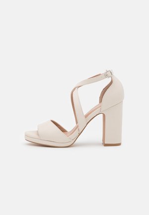 Beige højhælet sandal med tyk blokhæl, åben tå, krydsede ankelremme og en lille spænde lukning på en ensfarvet baggrund.