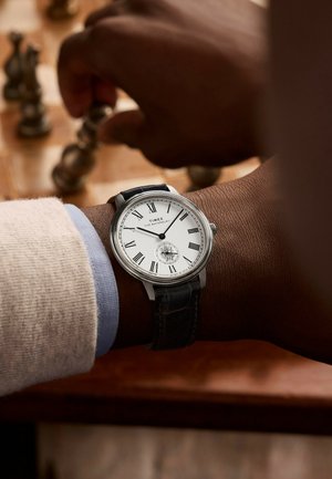 Montre Timex en argent avec un cadran blanc, des chiffres romains et un design squelettique ; bracelet en cuir noir ; main positionnant une pièce d'échecs.