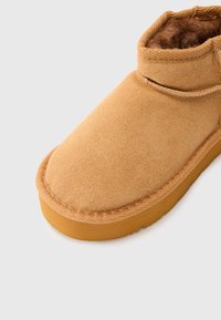 Botín de ante marrón con un suave forro de piel de peluche, puntera redonda y costuras decorativas. Cuenta con una suela de goma para durabilidad y agarre.