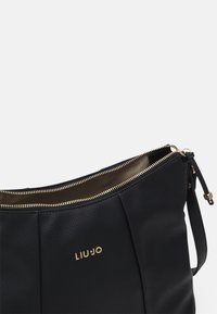 Borsa in pelle nera con finitura texturizzata, dotata di una cerniera dorata e logo. L'interno è beige, con una singola tracolla per il trasporto.