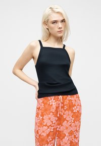 Jeune femme aux cheveux blonds portant un haut noir sans manches et un pantalon orange à motifs floraux, posant avec une main sur la hanche.