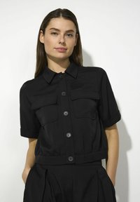Zwart shirt met korte mouwen, een gestructureerde kraag, twee borstzakken en knoopsluiting. Gemaakt van een gladde stof met een lichte glans.