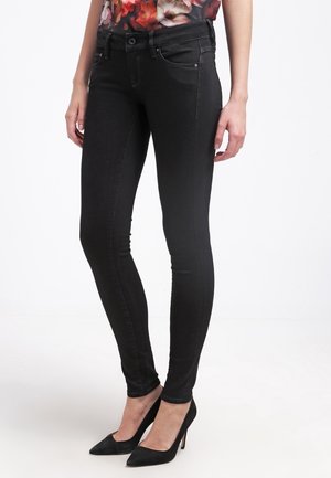 Femme portant un jean skinny noir et des talons aiguilles noirs à bout pointu, debout devant un fond blanc.