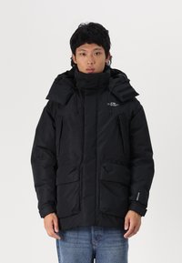 Sort polstret vinterjakke med høj krave, justerbar hætte, flere frontlommer og Velcro-detaljer. Glat tekstur, elegant design.