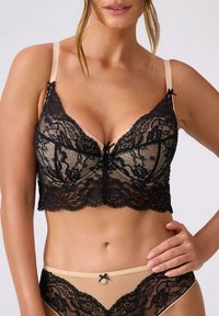 Kobieta w beżowej bieliźnie złożonej z misternie czarnej koronkowej aplikacji na biustonoszu i majtkach, stojąca z jedną ręką na biodrze na tle jednolitego tła.