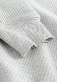 Világosszürke bordázott anyag részletes textúrával, behajtott mandzsettával. Az anyag puhanak és enyhén rugalmasnak tűnik.