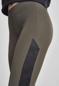 Alyvuogių spalvos legginsai su lygia tekstūra, šone turintys juodos spalvos kamufliažo rašto tinklinį intarpą vėdinimui ir stiliui.